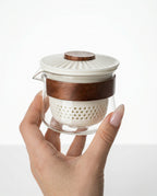 Hand holding a small glass infuser for gongfu ceremony with wooden accents on a white background