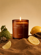 An amber glass jar candle labeled 'HIGH TEA BLACK TEA • LEMON • BERGAMOT' by the brand e sho, lit and centered on a sandy surface with lemon slices, a bergamot fruit, and dried botanicals around it against a warm beige background.