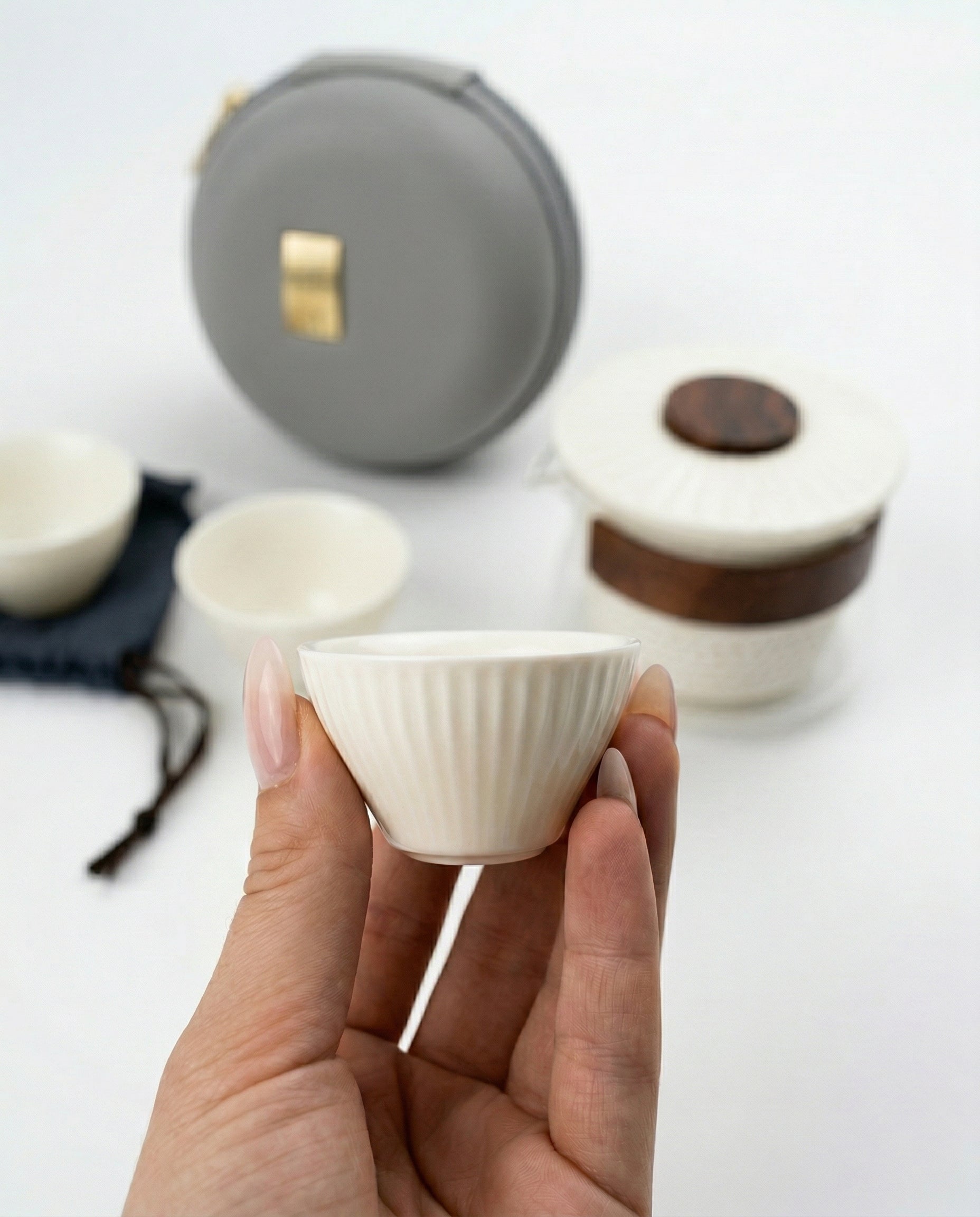 Person holding a small white ceramic cup with more cups and a gray case in the background.